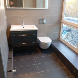 Bauelektro - Nowoczesna łazienka po remoncie: szare płytki, podwieszana toaleta, szafka z umywalką i lustro z podświetleniem LED. Widok z poziomu podłogi.