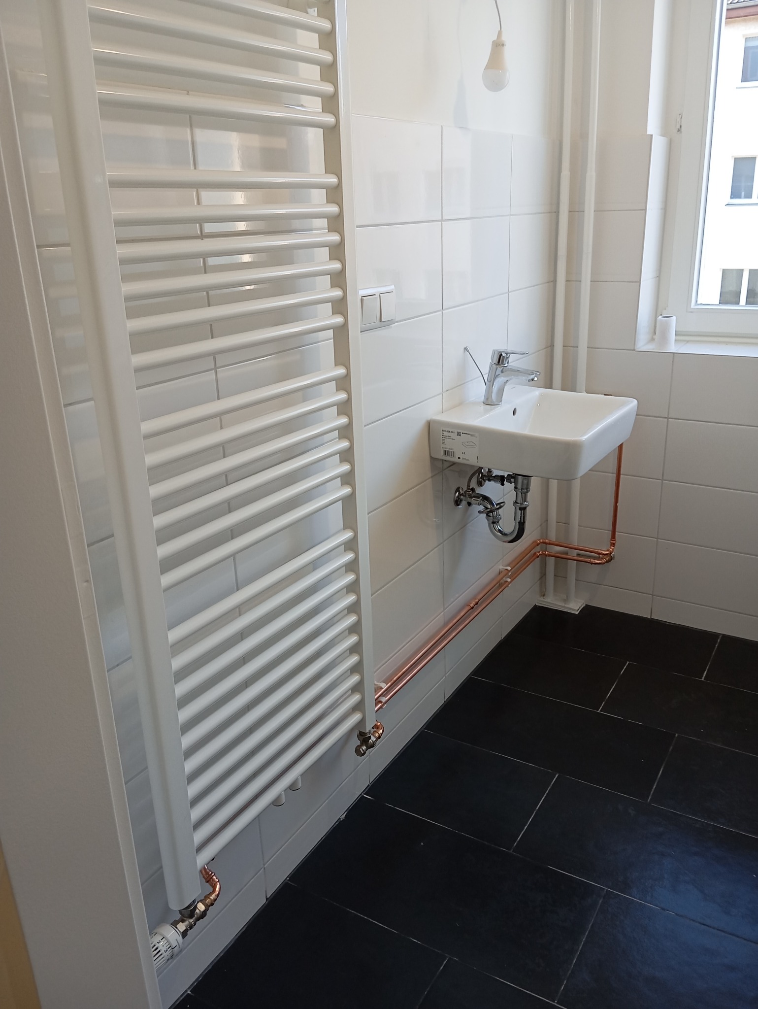 Biała umywalka z baterią i grzejnik drabinkowy na tle białych płytek w łazience z czarną podłogą. Widoczne miedziane rury instalacji wodnej.