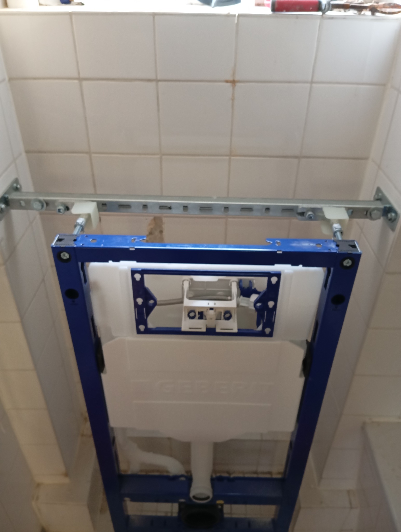 Stelaż podtynkowy Geberit Duofix zamontowany w łazience z białymi płytkami, widoczne elementy mocujące do ściany. Ujęcie z dołu, narzędzia na górze kadru.