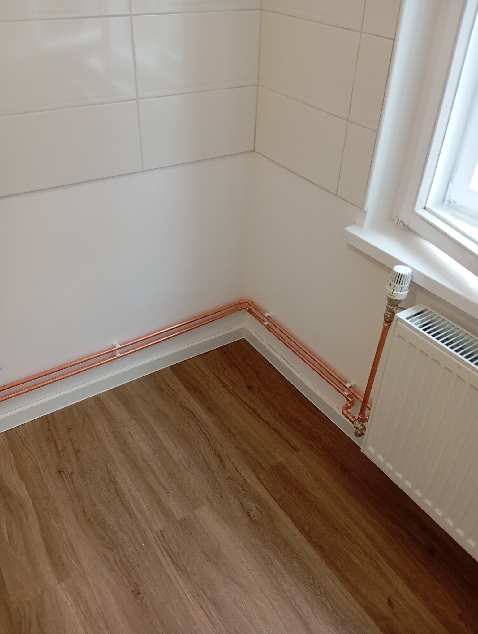 Miedziane rury grzewcze zamontowane wzdłuż ściany z białymi płytkami i drewnianą podłogą, widoczne przyłącze do grzejnika pod oknem.
