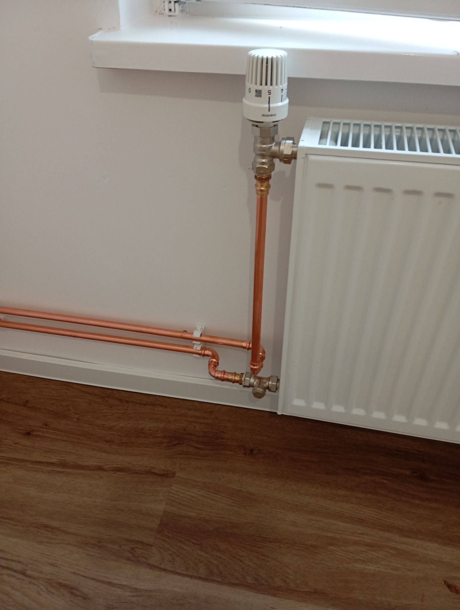 Biały grzejnik z termostatem i miedzianymi rurami, zamontowany przy białej ścianie i oknie, na tle podłogi z paneli imitujących drewno.