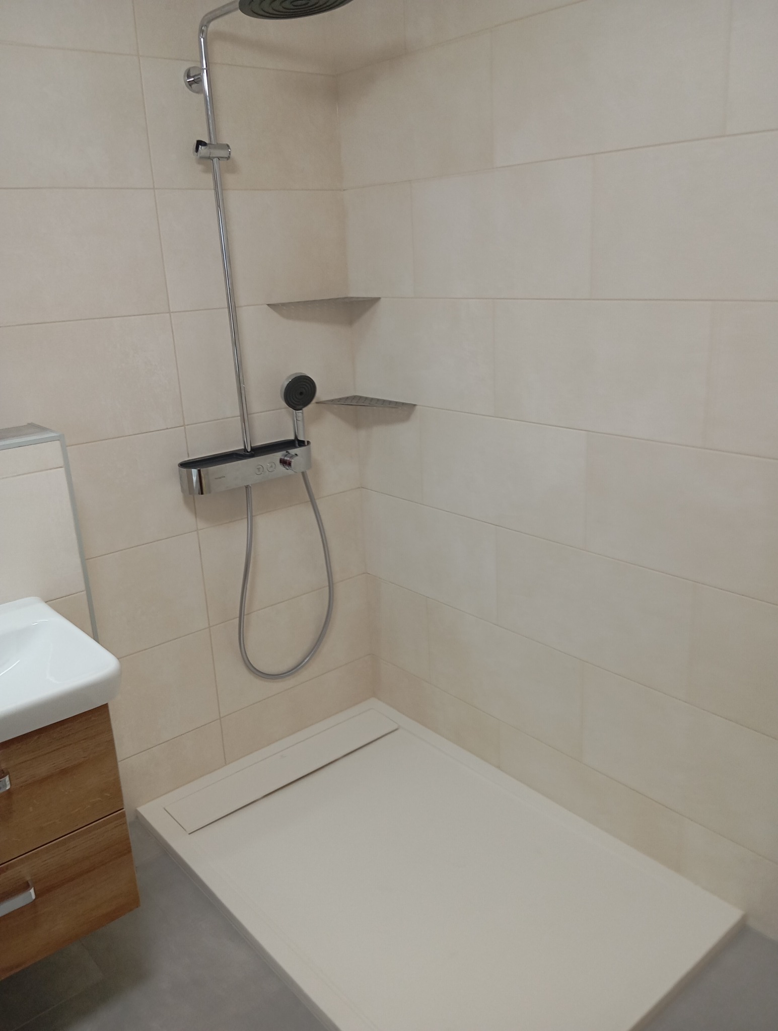 Nowoczesna kabina prysznicowa z deszczownicą i termostatem, wykończona beżowymi płytkami. Umywalka z szafką częściowo widoczna po lewej stronie. Minimalistyczny design.