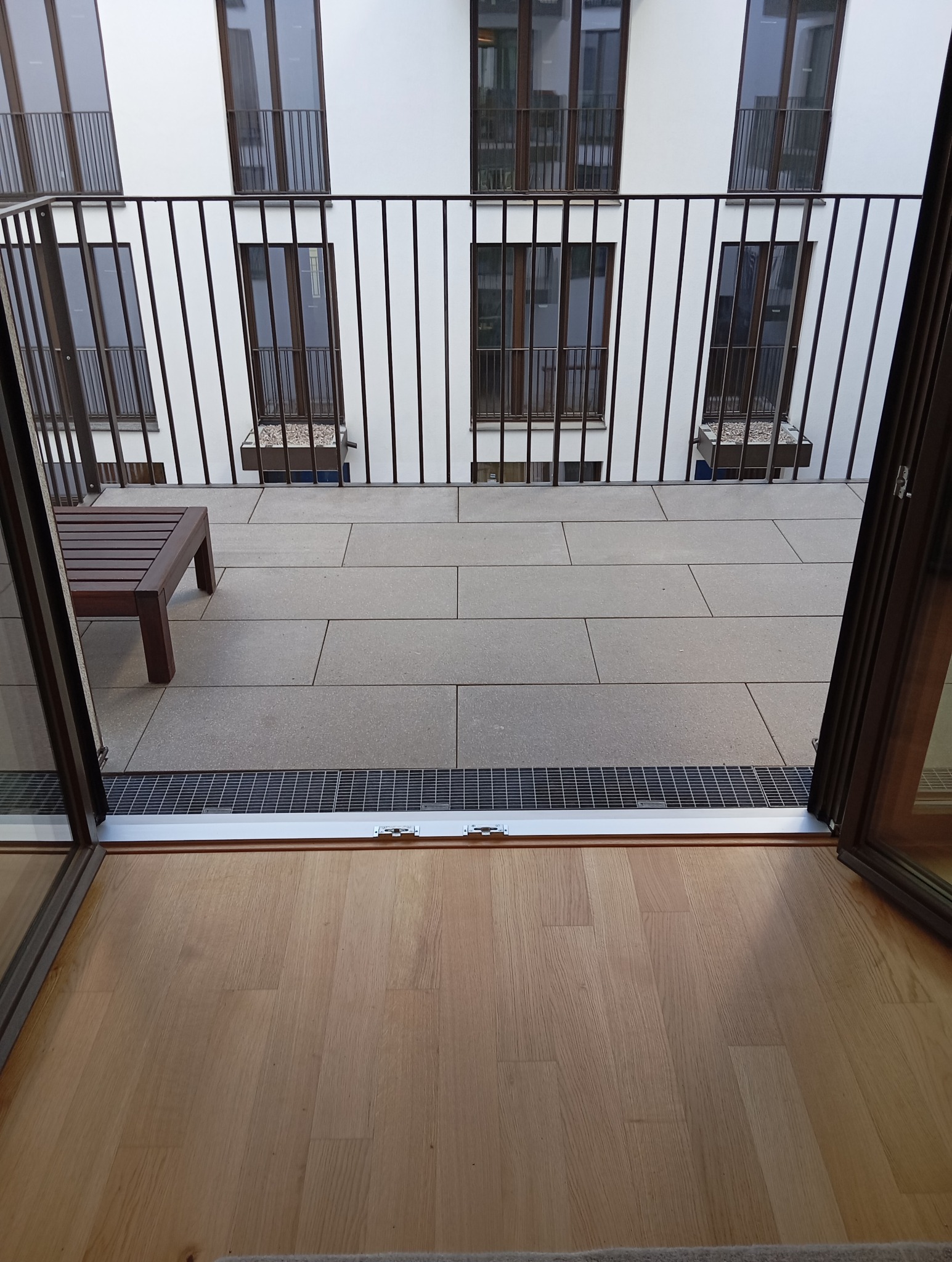 Nowoczesny balkon z szarymi płytami, metalową balustradą i drewnianą ławką, widok z wnętrza przez otwarte drzwi balkonowe. Minimalistyczny design.