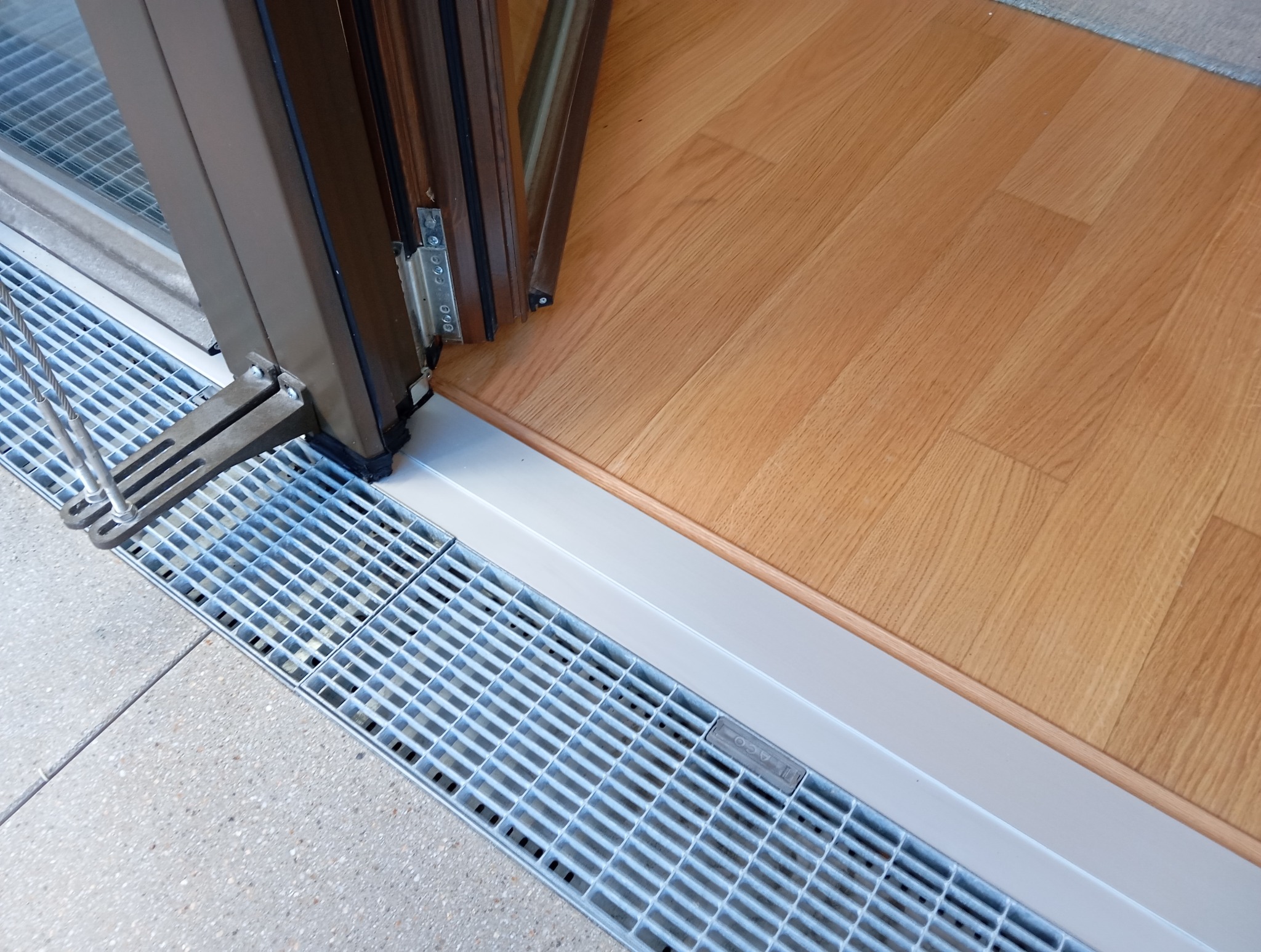 Otwarta, brązowa rama drzwi balkonowych z widocznym systemem odprowadzania wody. Podłoga z paneli drewnianych i szara, betonowa posadzka na zewnątrz.