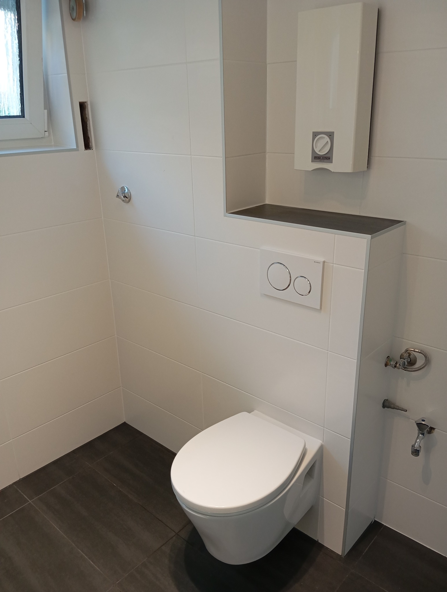 Nowoczesna toaleta podwieszana z białymi płytkami i ciemną podłogą. Widoczny bojler nad zabudową. Minimalistyczny design, czyste linie, schludne wykończenie.