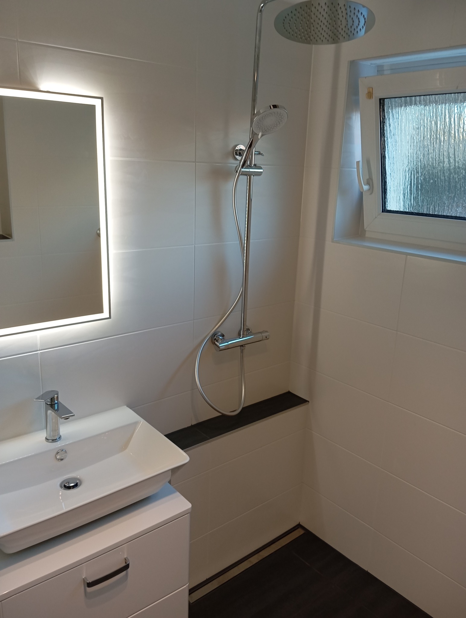 Nowoczesna łazienka z białą armaturą, kabiną prysznicową typu walk-in i podświetlanym lustrem. Minimalistyczny design z ciemnymi akcentami na podłodze i półce.