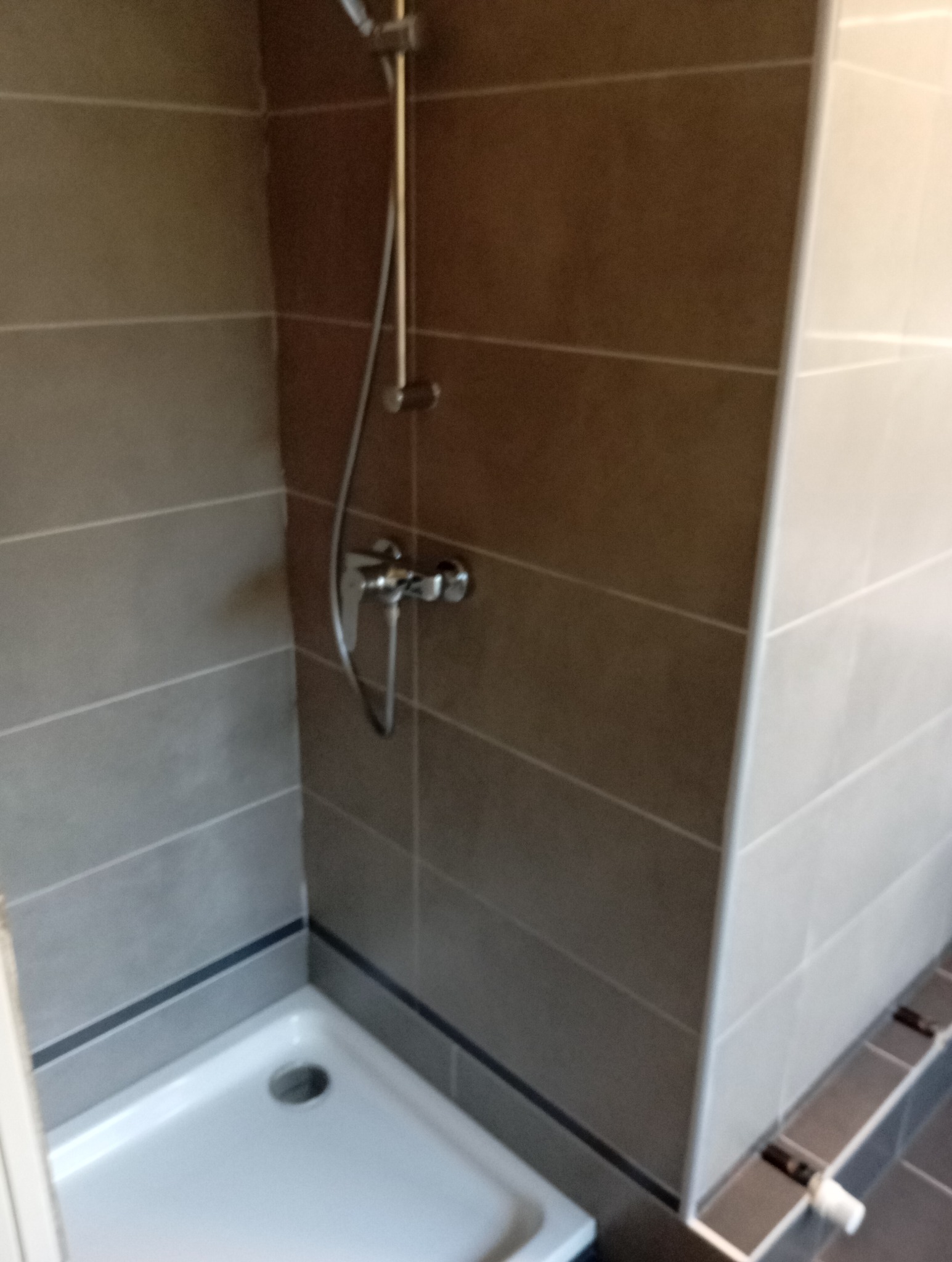 Wykończona kabina prysznicowa z szarymi płytkami, chromowaną armaturą i białym brodzikiem. Widoczne detale wykończenia i minimalistyczny design.