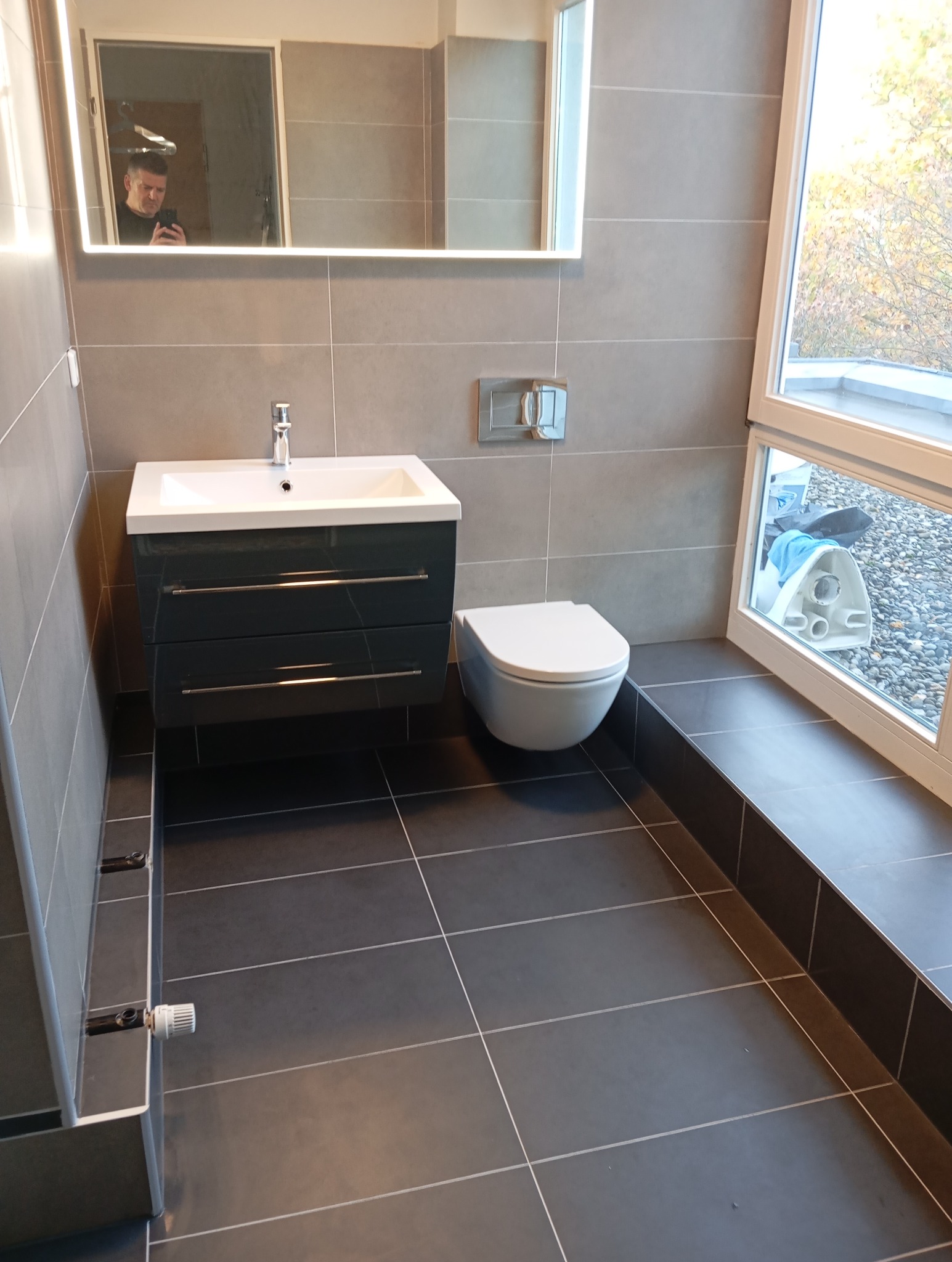 Nowoczesna łazienka po remoncie: szare płytki, podwieszana toaleta, szafka z umywalką i lustro z podświetleniem LED. Widok z poziomu podłogi.