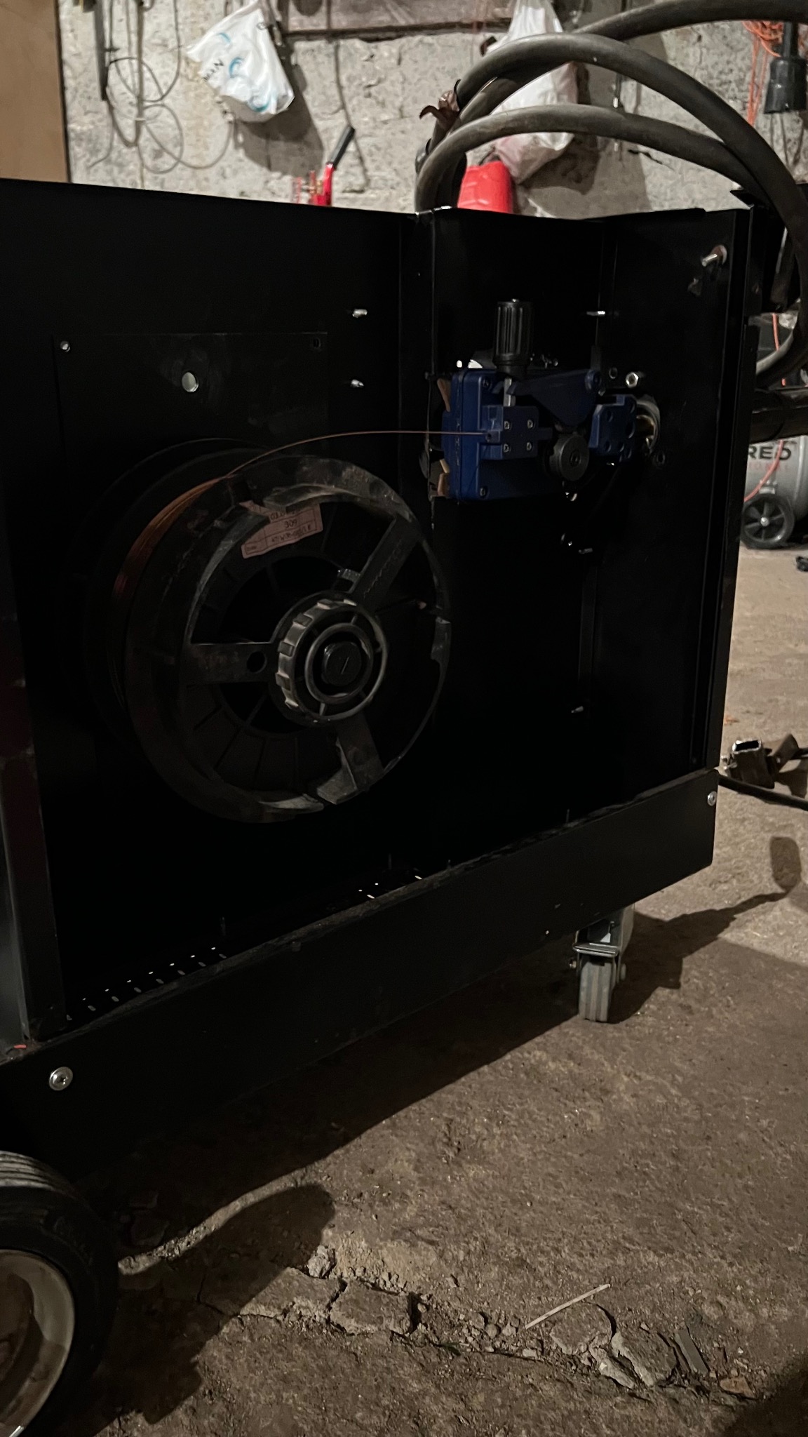 Wnętrze spawarki z widoczną szpulą drutu spawalniczego i mechanizmem podającym, umieszczone w czarnej obudowie na kółkach.