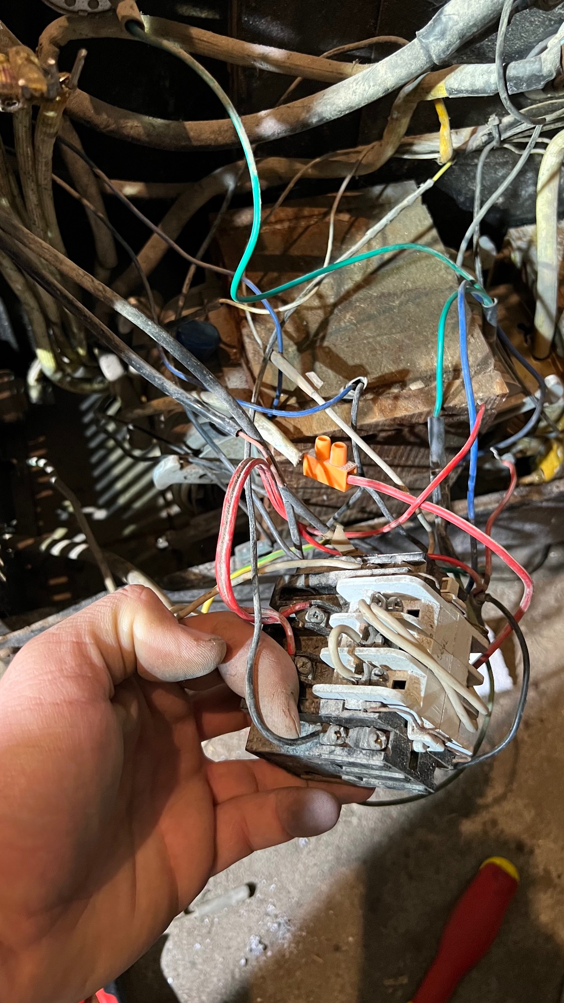 Brudny stycznik elektryczny trzymany w dłoni na tle splątanych przewodów i kabli. Widoczny pomarańczowy konektor. Na podłodze leży śrubokręt z czerwoną rękojeścią. Zdjęcie wykonane przed naprawą.