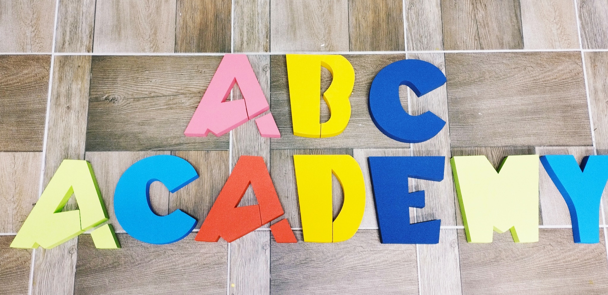 Kolorowe, piankowe litery układające się w napis 'ABC Academy' na drewnopodobnej podłodze w Wągrowcu. Litera A jest różowa i pęknięta, B żółta, C niebieska, reszta liter w różnych kolorach.