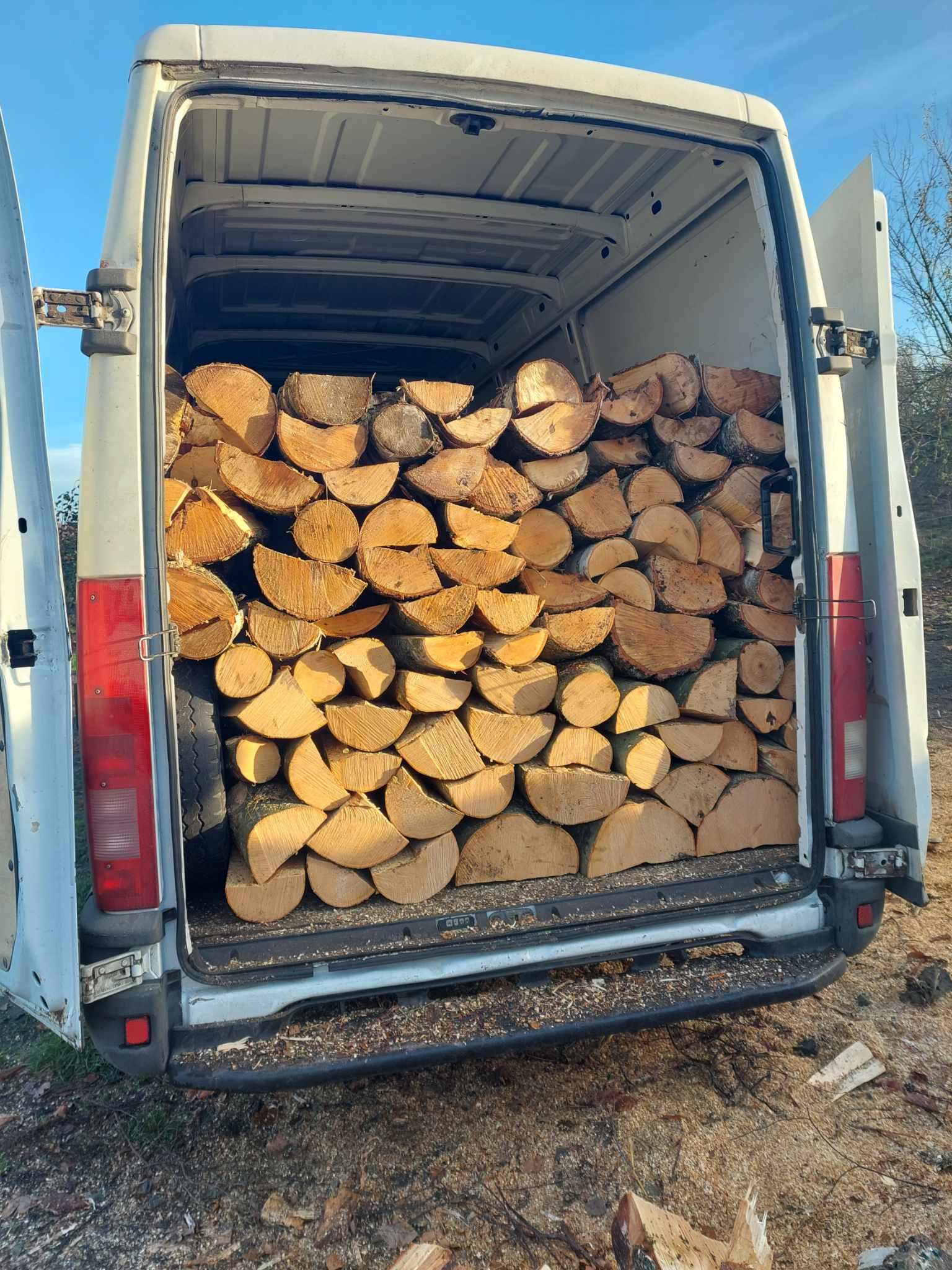 Pełny bus z otwartymi drzwiami załadowany pociętymi kawałkami drewna na opał, widoczne opony w przestrzeni ładunkowej.