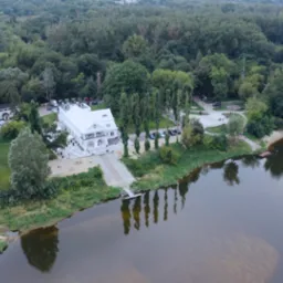 Panoramiczny widok z lotu ptaka na biały budynek z tarasem nad rzeką, otoczony zielenią, z zaparkowanymi samochodami i przygotowanym terenem, prawdopodobnie pod imprezę.
