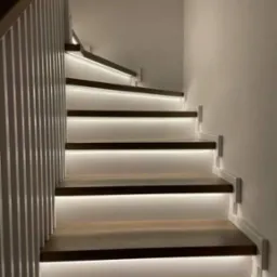 Nowoczesne schody z podświetleniem LED pod stopniami, biała balustrada z pionowymi szczebelkami, ujęcie od dołu.