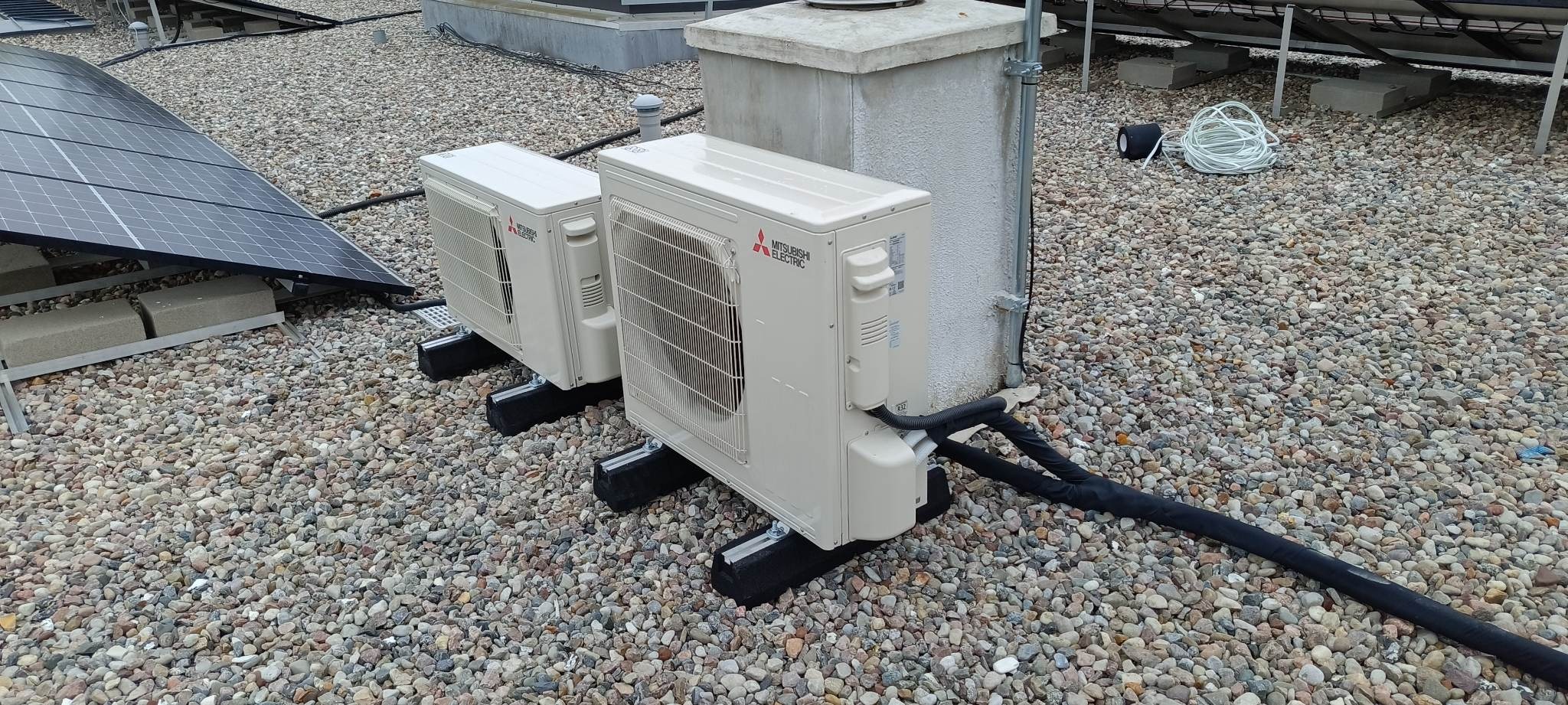 Dwa zewnętrzne moduły klimatyzacji Mitsubishi Electric na dachu pokrytym żwirem, z widocznymi panelami fotowoltaicznymi w tle i przewodami w izolacji.