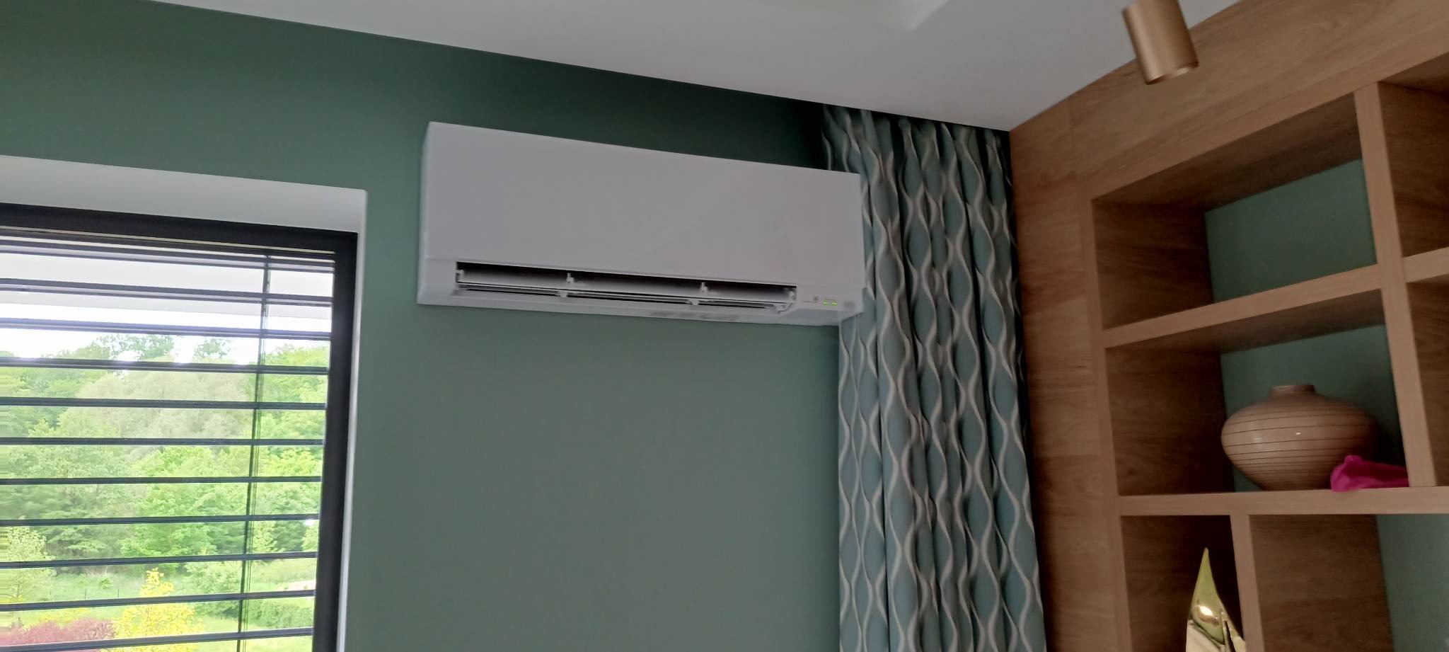 Biała jednostka wewnętrzna klimatyzacji zamontowana na zielonej ścianie w nowoczesnym wnętrzu, obok okna z żaluzjami i dekoracyjnych zasłon oraz drewnianego regału z ozdobną wazą.