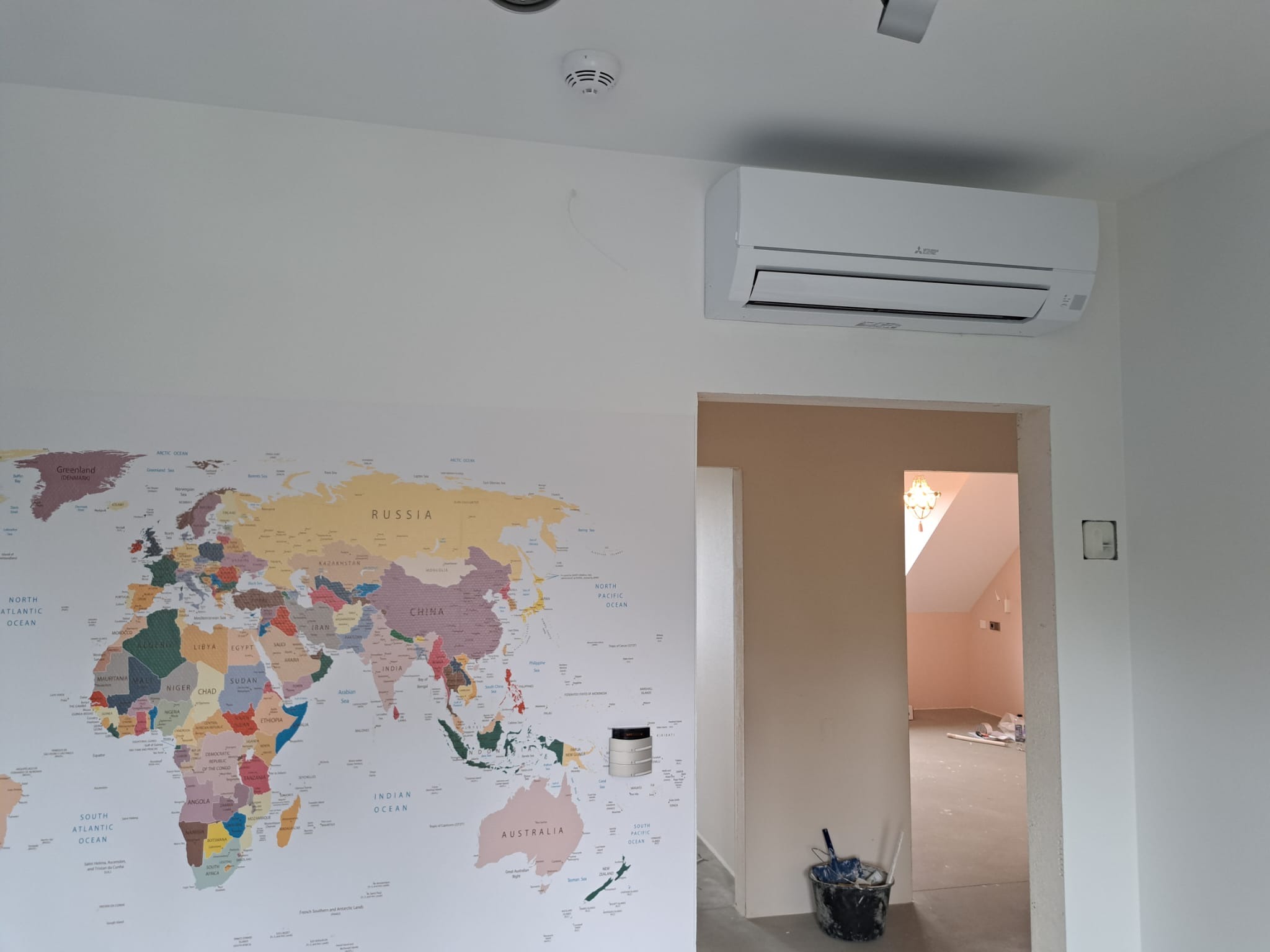 Biała klimatyzacja Mitsubishi zamontowana na ścianie w pokoju z kolorową mapą świata, widoczny czujnik dymu na suficie i otwarte drzwi do remontowanego pomieszczenia.