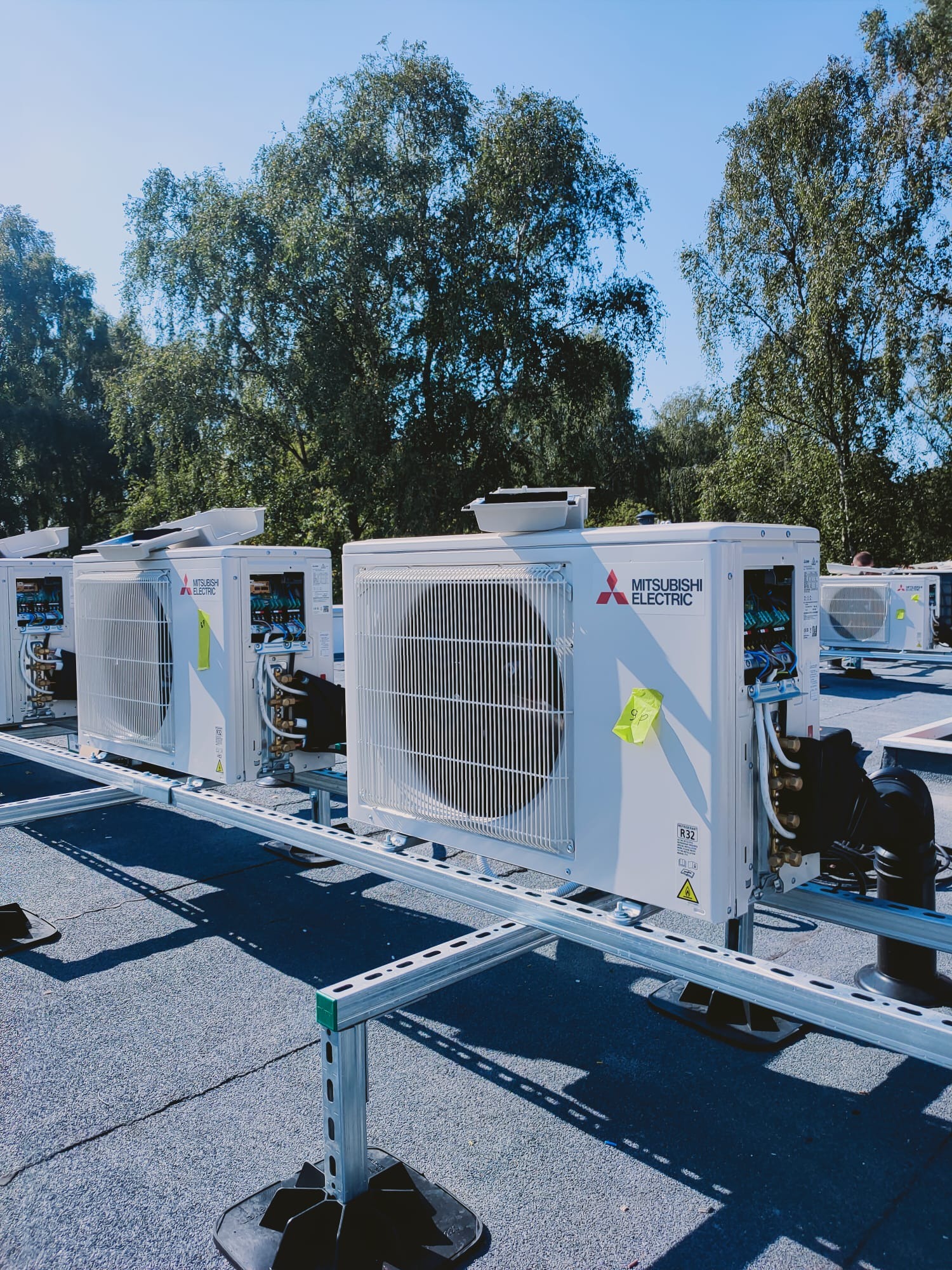 Rząd białych jednostek zewnętrznych klimatyzacji Mitsubishi Electric zamontowanych na dachu, widoczne przewody i naklejka z napisem '90'.