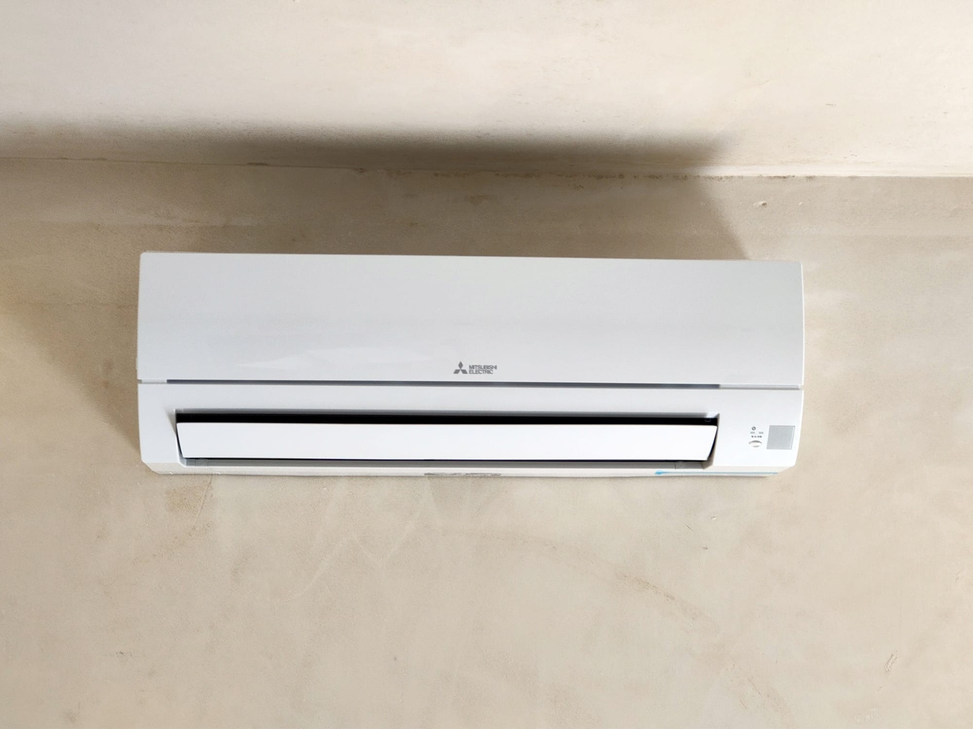 Biała jednostka klimatyzacji Mitsubishi Electric zamontowana na beżowej ścianie z widoczną fakturą. Klapka nawiewu jest otwarta.