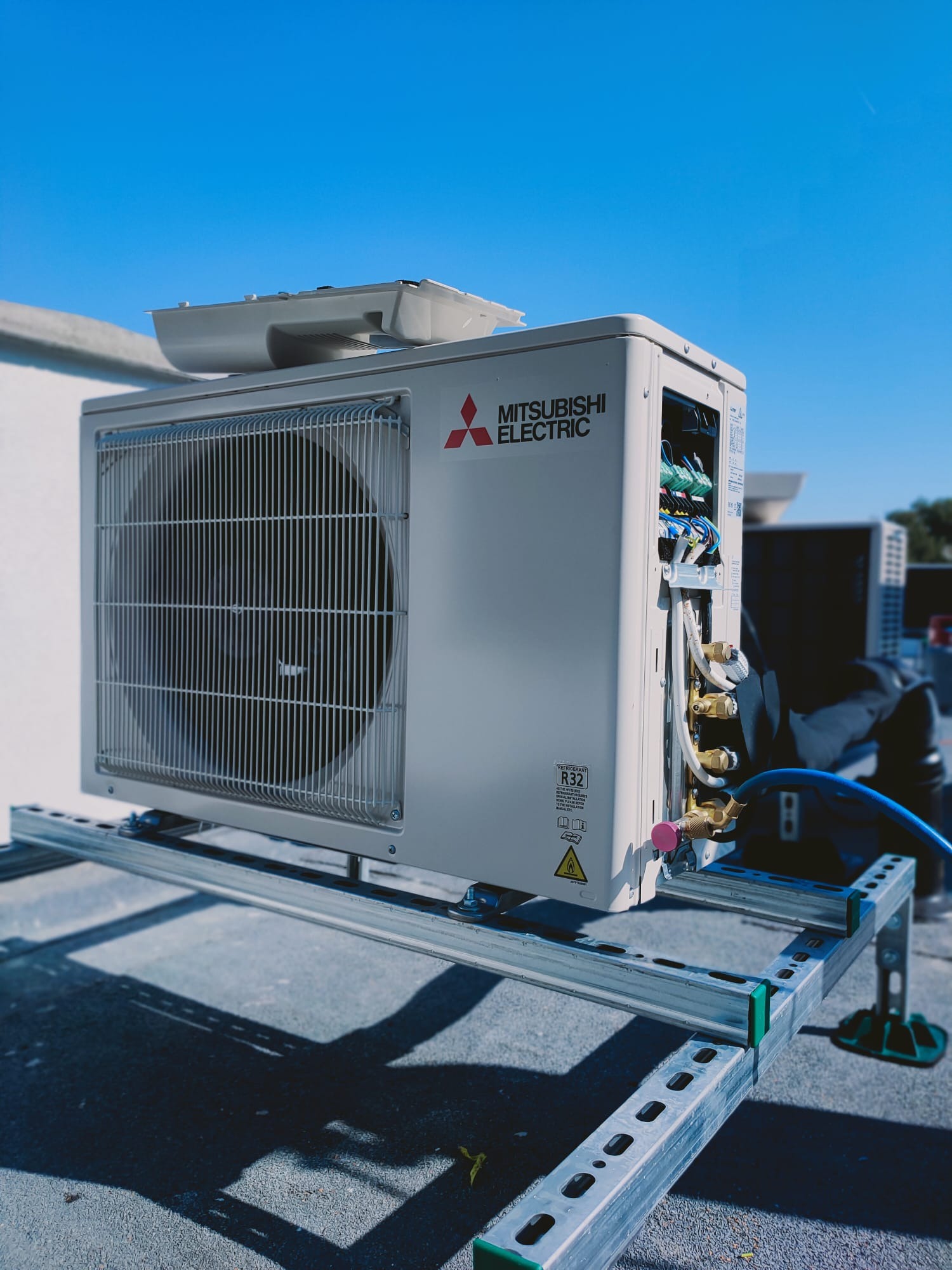 Zewnętrzna jednostka klimatyzacji Mitsubishi Electric na metalowej konstrukcji wsporczej z widocznymi przewodami i niebieskim wężem serwisowym, na dachu budynku, w słoneczny dzień.