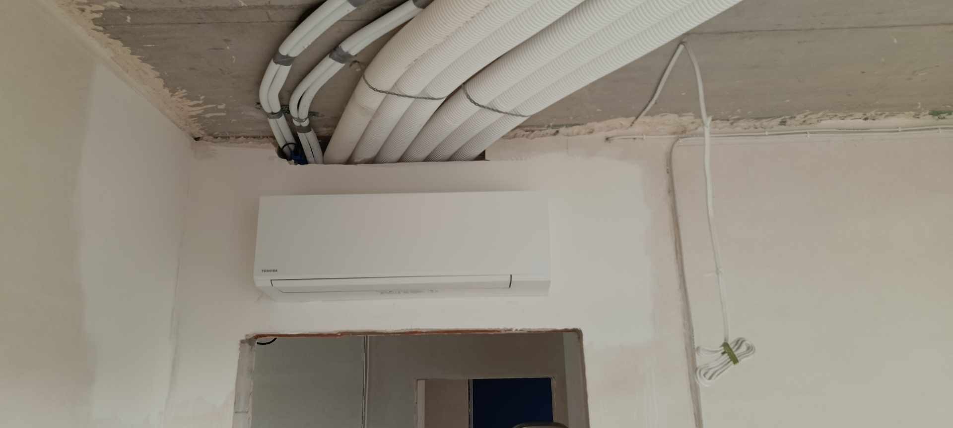 Biały klimatyzator Toshiba zamontowany na ścianie w surowym wnętrzu, widoczne rury instalacyjne pod sufitem i otwór drzwiowy w tle.