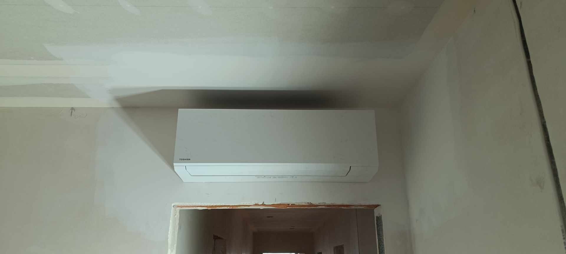 Biała jednostka wewnętrzna klimatyzacji Toshiba zamontowana na ścianie w surowym stanie, widoczne przygotowania do wykończenia wnętrza, ściany w szpachli, rama drzwi bez ościeżnicy.