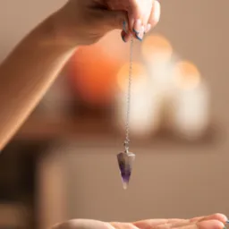 Kobieta trzyma wahadełko z fioletowego kamienia na srebrnym łańcuszku nad otwartą dłonią, w tle rozmyte świece i pomarańczowa kula.