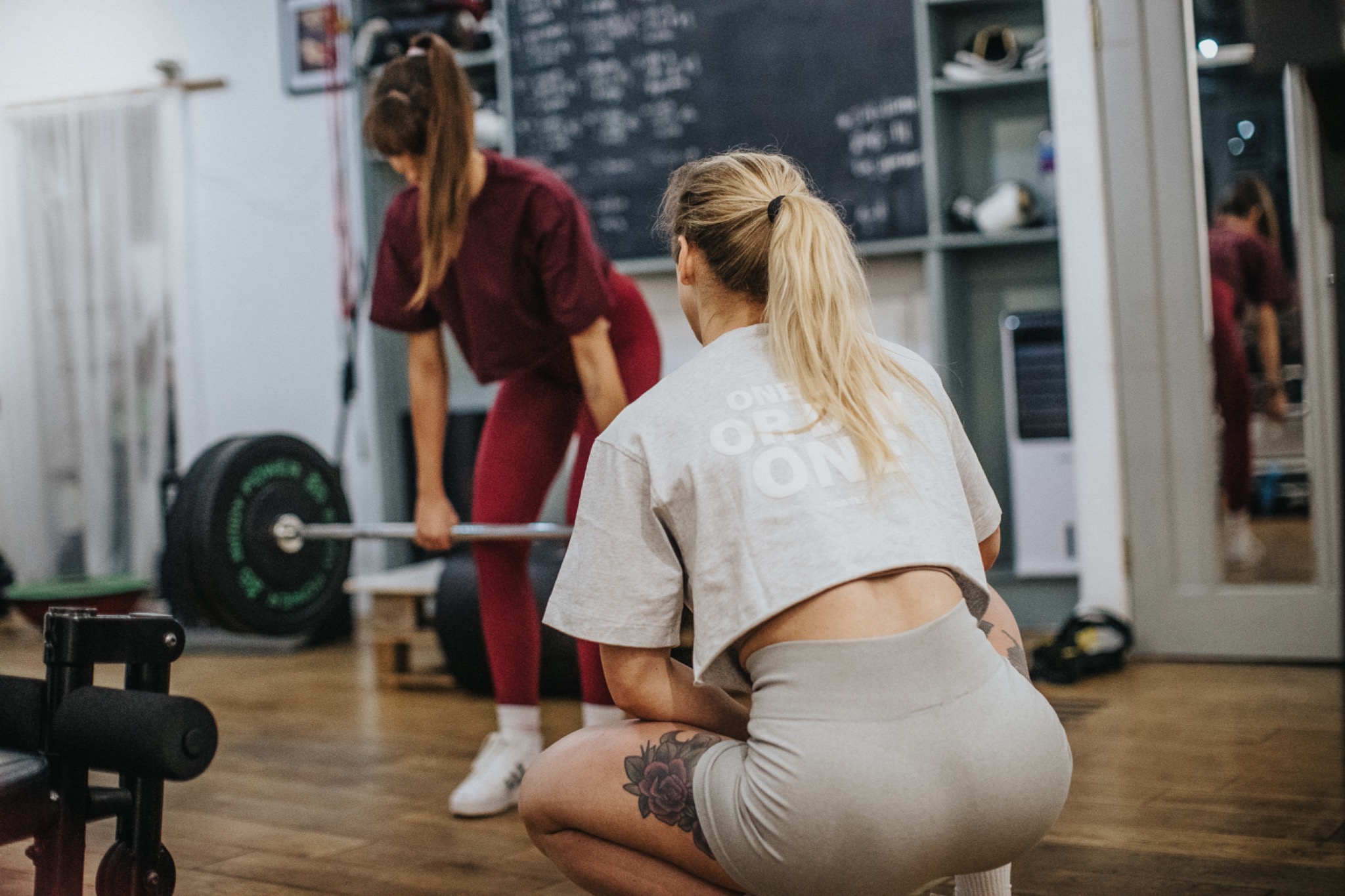 Kobieta w sportowym stroju kuca, obserwując drugą osobę podnoszącą sztangę w siłowni z drewnianą podłogą i tablicą w tle.