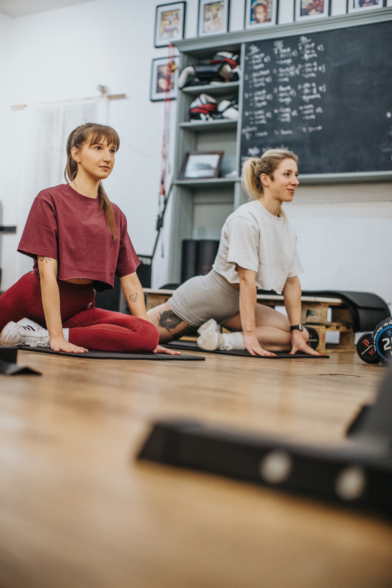 Dwie kobiety wykonują ćwiczenia rozciągające w jasnym pomieszczeniu z tablicą treningową w tle; jedna w bordowym stroju, druga w szarym, obie na matach do ćwiczeń.