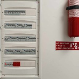 Mika'EL - Otwarta skrzynka z bezpiecznikami na białej ścianie, obok gaśnica do sprzętu komputerowego i tabliczka informacyjna