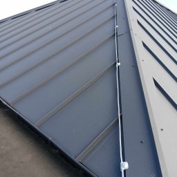 Mika'EL - Szary dach z blachy trapezowej z zainstalowaną instalacją odgromową, widoczne uchwyty mocujące przewód odprowadzający.