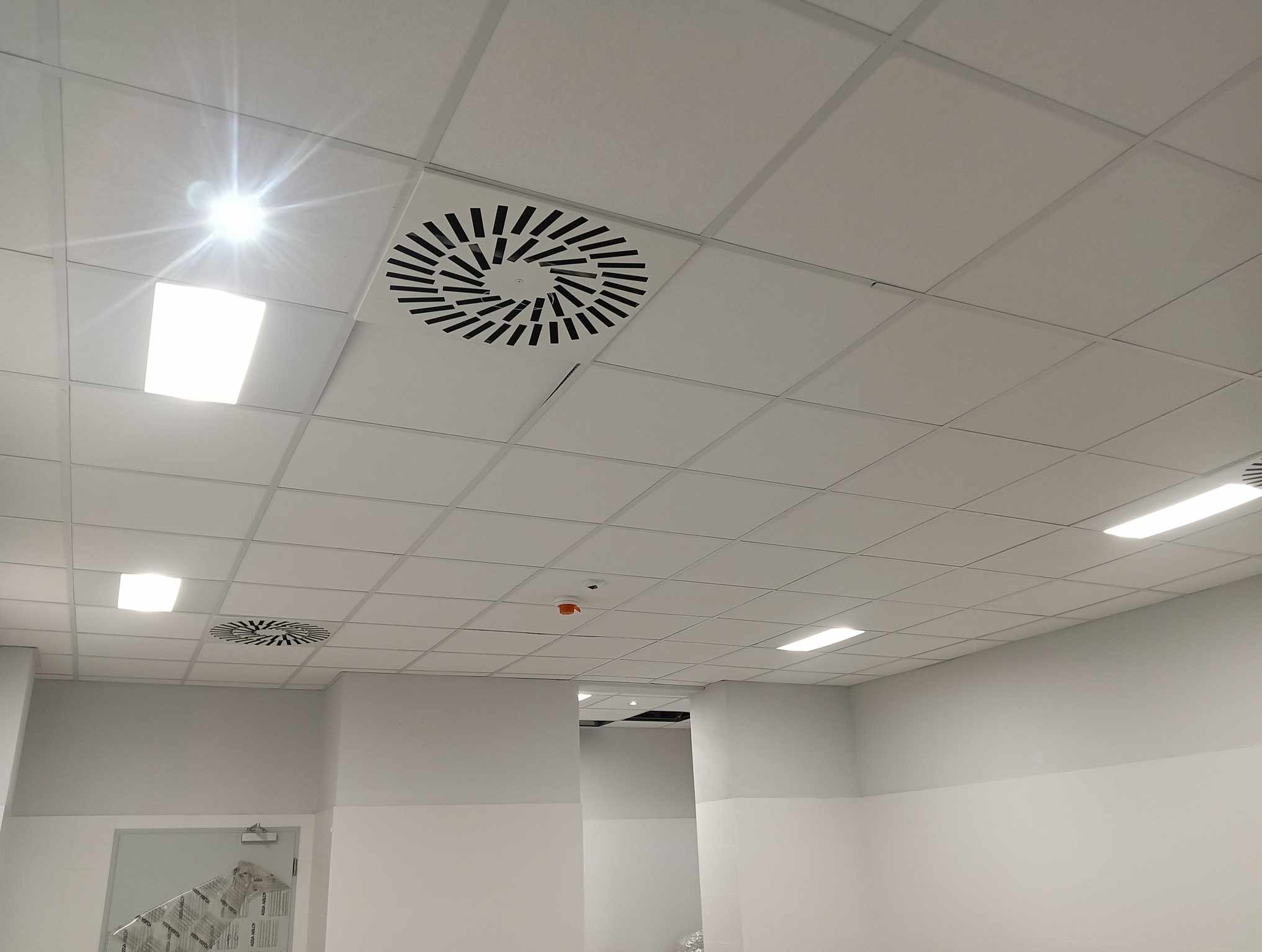 Sufit podwieszany z kwadratowych paneli z widocznymi lampami LED i okrągłymi kratkami wentylacyjnymi, w tle widoczny czujnik dymu i fragment korytarza.