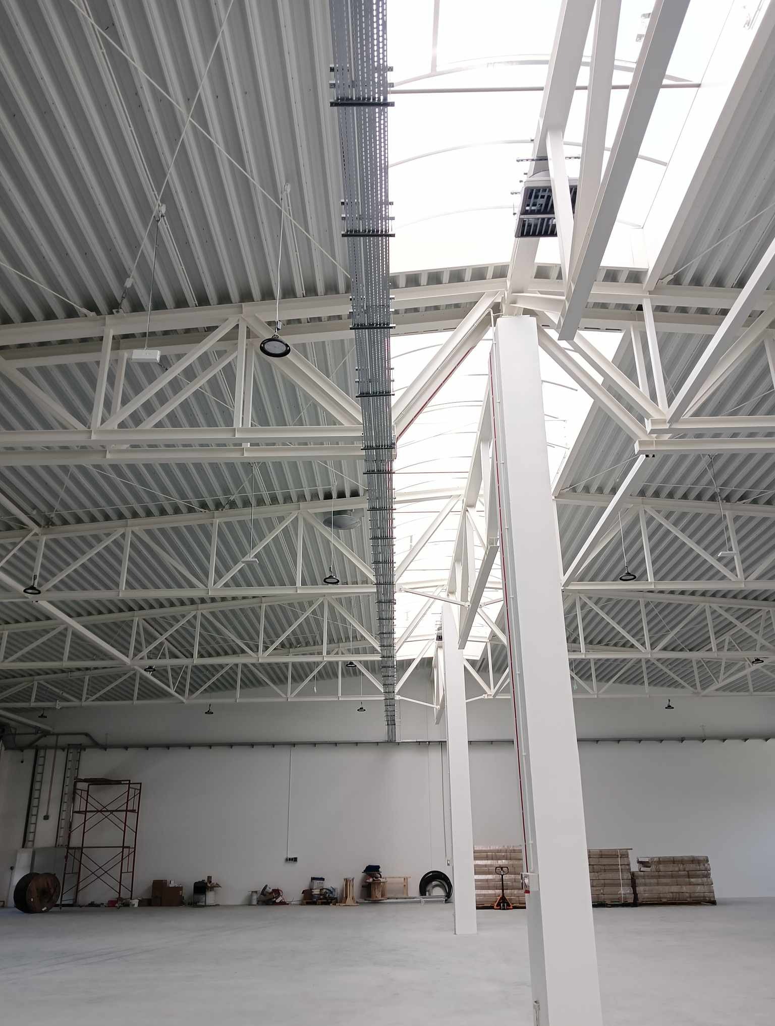 Wnętrze hali magazynowej z widoczną konstrukcją dachu, oświetleniem LED, instalacjami biegnącymi pod sufitem oraz elementami wyposażenia na podłodze.
