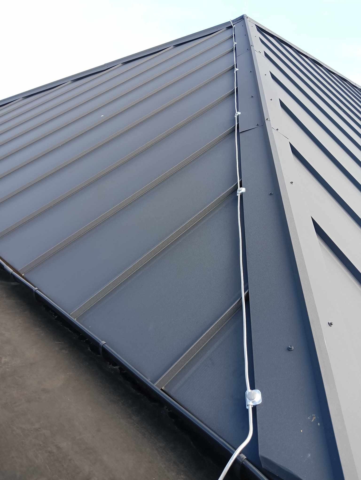 Szary dach z blachy trapezowej z zainstalowaną instalacją odgromową, widoczne uchwyty mocujące przewód odprowadzający.