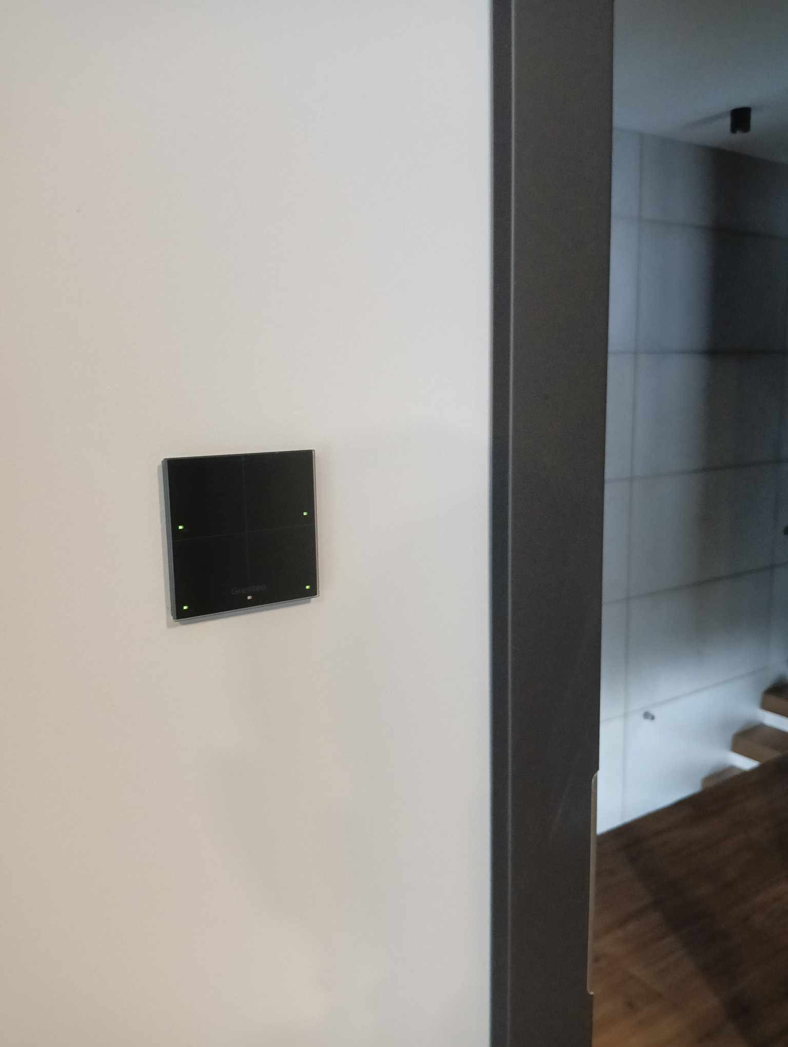 Czarny panel sterowania z zielonymi diodami LED zamontowany na białej ścianie, widok z boku przez szarą futrynę drzwi.