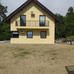 Żółty dom jednorodzinny z balkonem, czarnymi oknami i jednostkami klimatyzacji na elewacji; w tle widoczne drzewa i przygotowania do malowania elewacji.