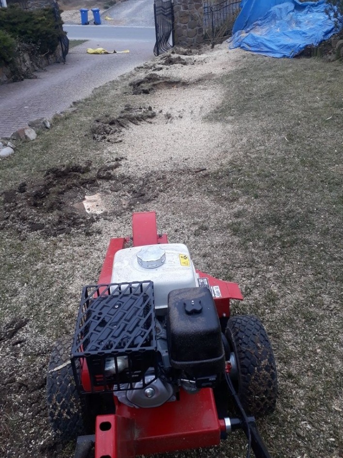 Czerwona rębarka do gałęzi na trawniku po wycince, z widocznymi śladami pracy i świeżymi trocinami. W tle brama i droga.