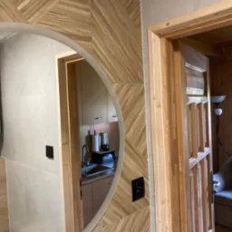 Wykończona łazienka z drewnopodobnymi płytkami ułożonymi w jodełkę na ścianie, okrągłe lustro z podświetleniem LED odbijające fragment kuchni, oraz drewniane drzwi z ościeżnicą.