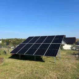 Panel słoneczny na metalowej konstrukcji, ustawiony na trawniku przed domem z ciemnym dachem, widoczny błękit nieba.