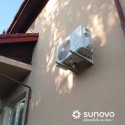 Biała jednostka zewnętrzna pompy ciepła marki Rotenso, zamontowana na beżowej ścianie domu, widoczna z boku w promieniach słońca.