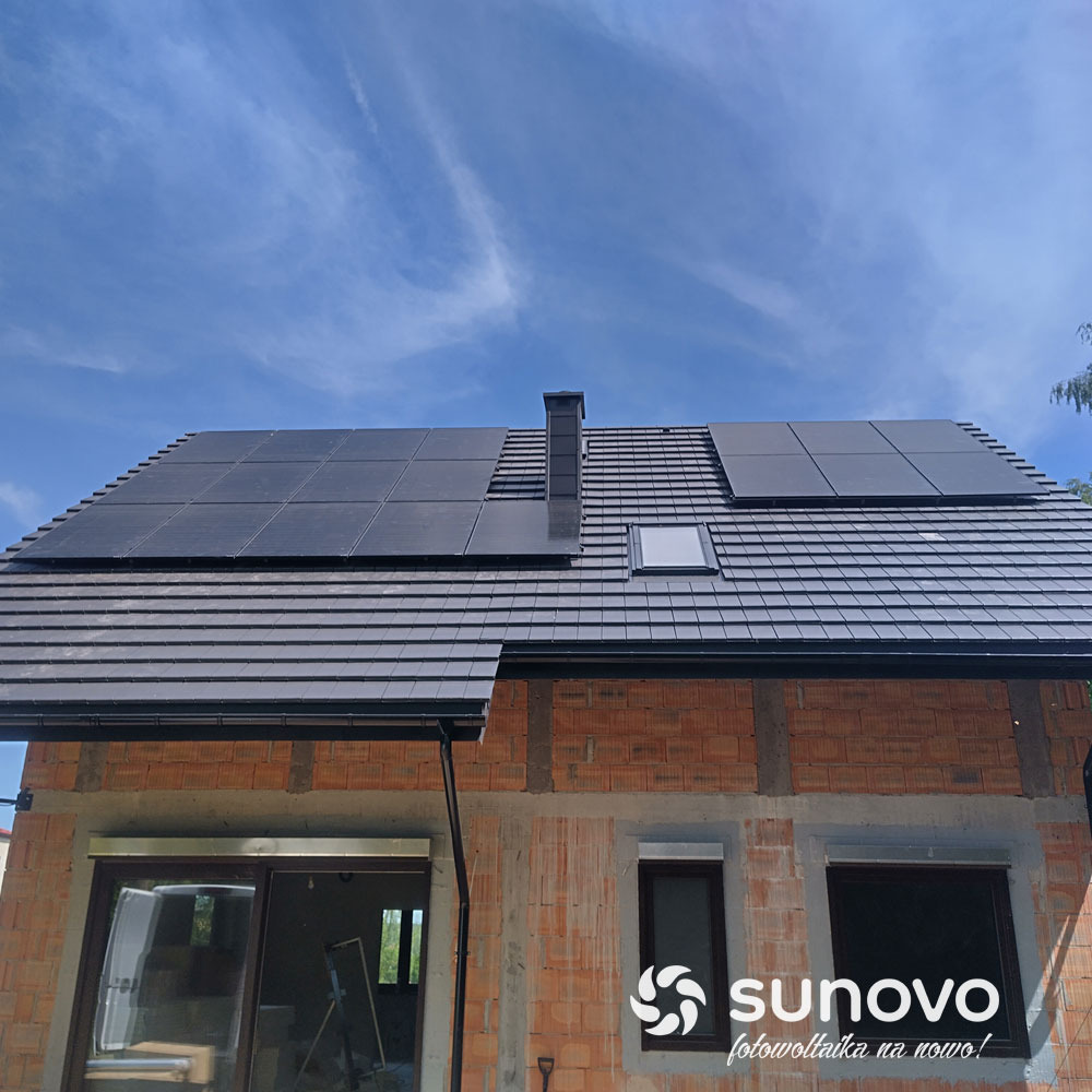 Częściowo otynkowany dom z czerwoną cegłą z zainstalowanymi panelami słonecznymi na dachu pokrytym ciemną dachówką; widoczne okno dachowe i komin; logo firmy w prawym dolnym rogu.