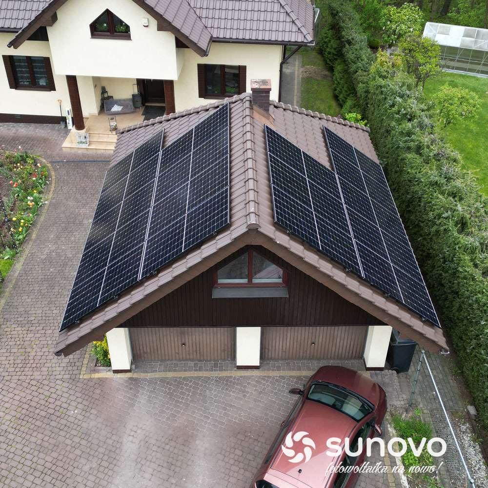 Widok z góry na dom z brązowym dachem, na którym zamontowano panele słoneczne; na podjeździe stoi bordowy samochód z logo firmy Sunovo.