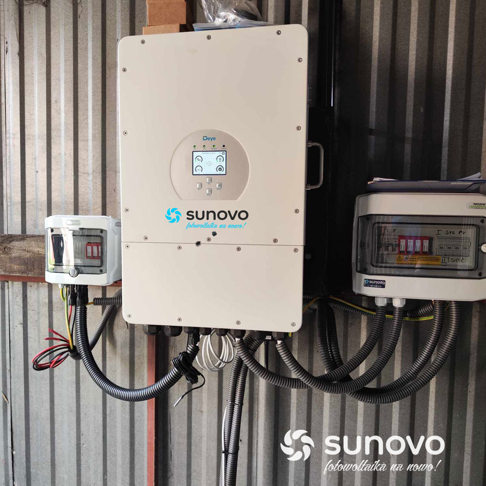 Zamontowany inwerter Sunovo z wyświetlaczem i skrzynkami zabezpieczeń na ścianie z blachy falistej. Widoczne przewody w peszlach.