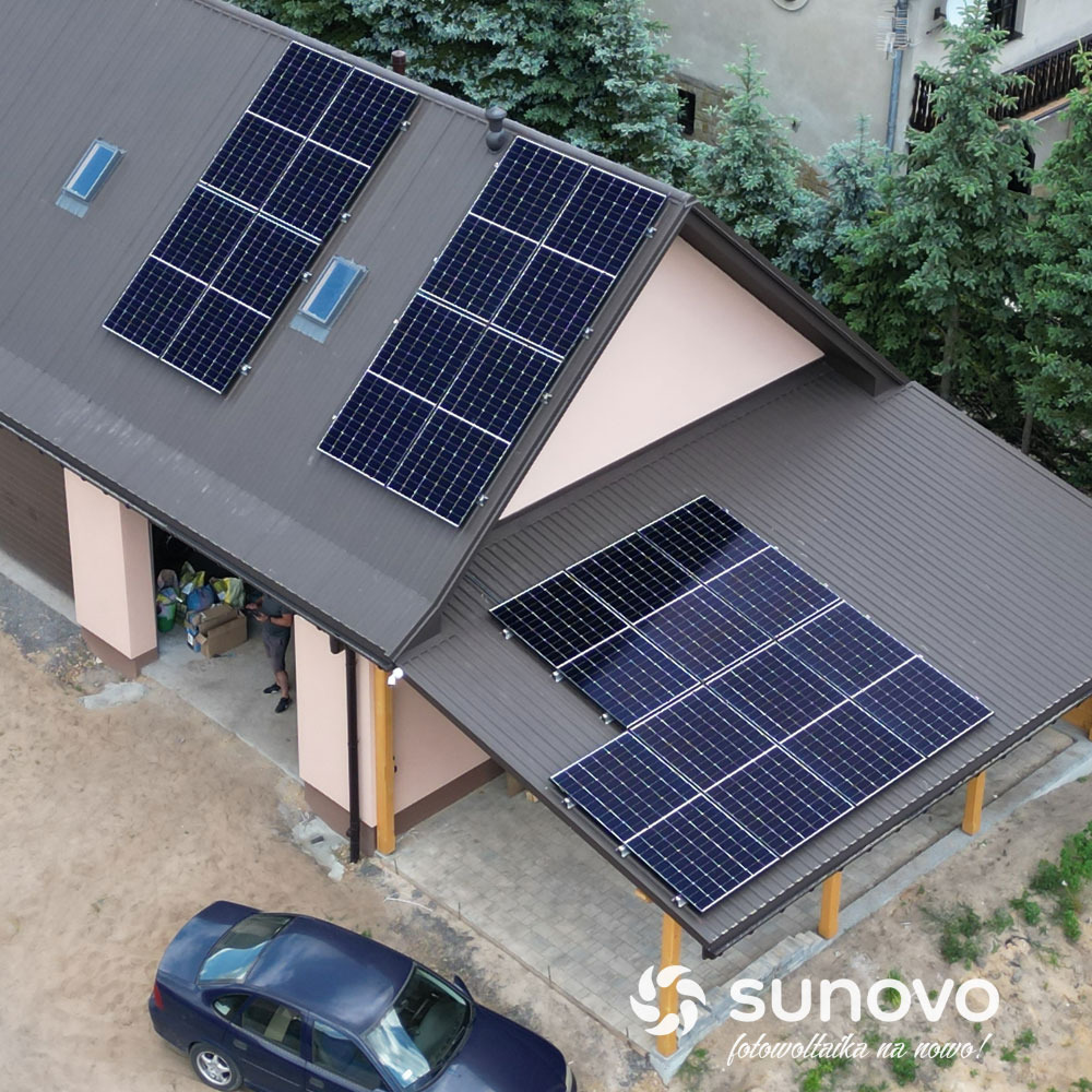 Instalacja paneli słonecznych na dachu domu z ciemnej blachy, widok z góry, błękitny samochód i logo firmy Sunovo w rogu zdjęcia.