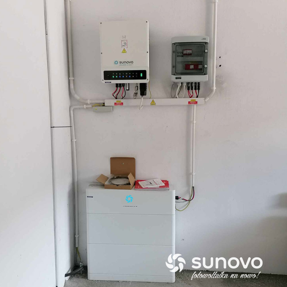 Biały magazyn energii Sunovo z otwartym kartonem akcesoriów na górze, podłączony do falownika i skrzynki bezpiecznikowej na białej ścianie.