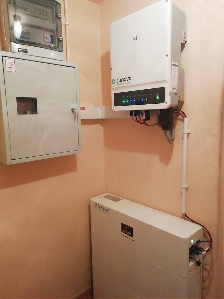 Instalacja domowa: biały falownik Sunovo z widocznymi diodami LED, obok skrzynka elektryczna z kluczykiem i biały magazyn energii Sunovo encor w rogu pomieszczenia o beżowych ścianach.