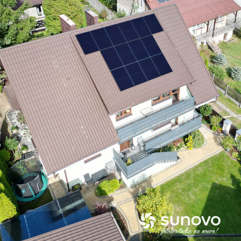 Widok z góry na dom z brązowym dachem i panelami fotowoltaicznymi ułożonymi w kształcie litery 'T', z widocznym ogrodem, trampoliną i logo firmy Sunovo w rogu.