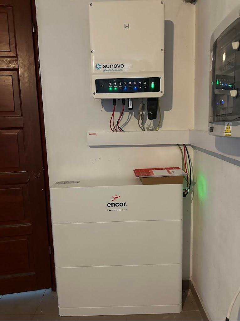 Biały inwerter Sunovo z zielonymi diodami LED i akumulator Encor zamontowane na ścianie wewnątrz pomieszczenia, obok brązowych drzwi.