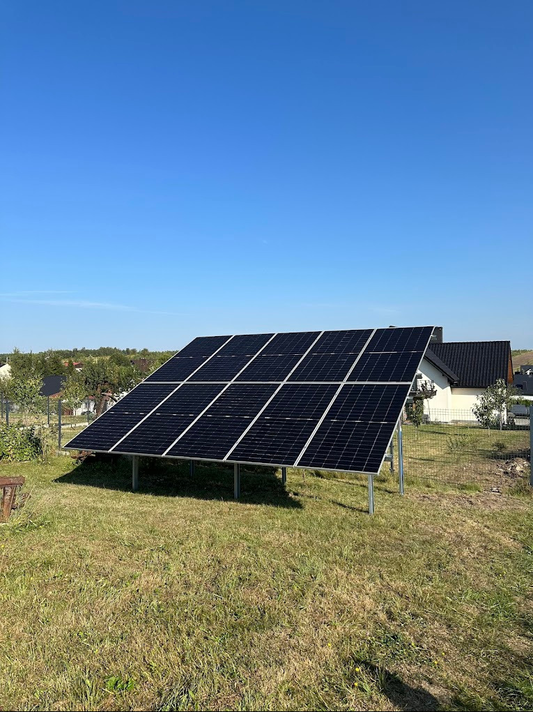 Panel słoneczny na metalowej konstrukcji, ustawiony na trawniku przed domem z ciemnym dachem, widoczny błękit nieba.
