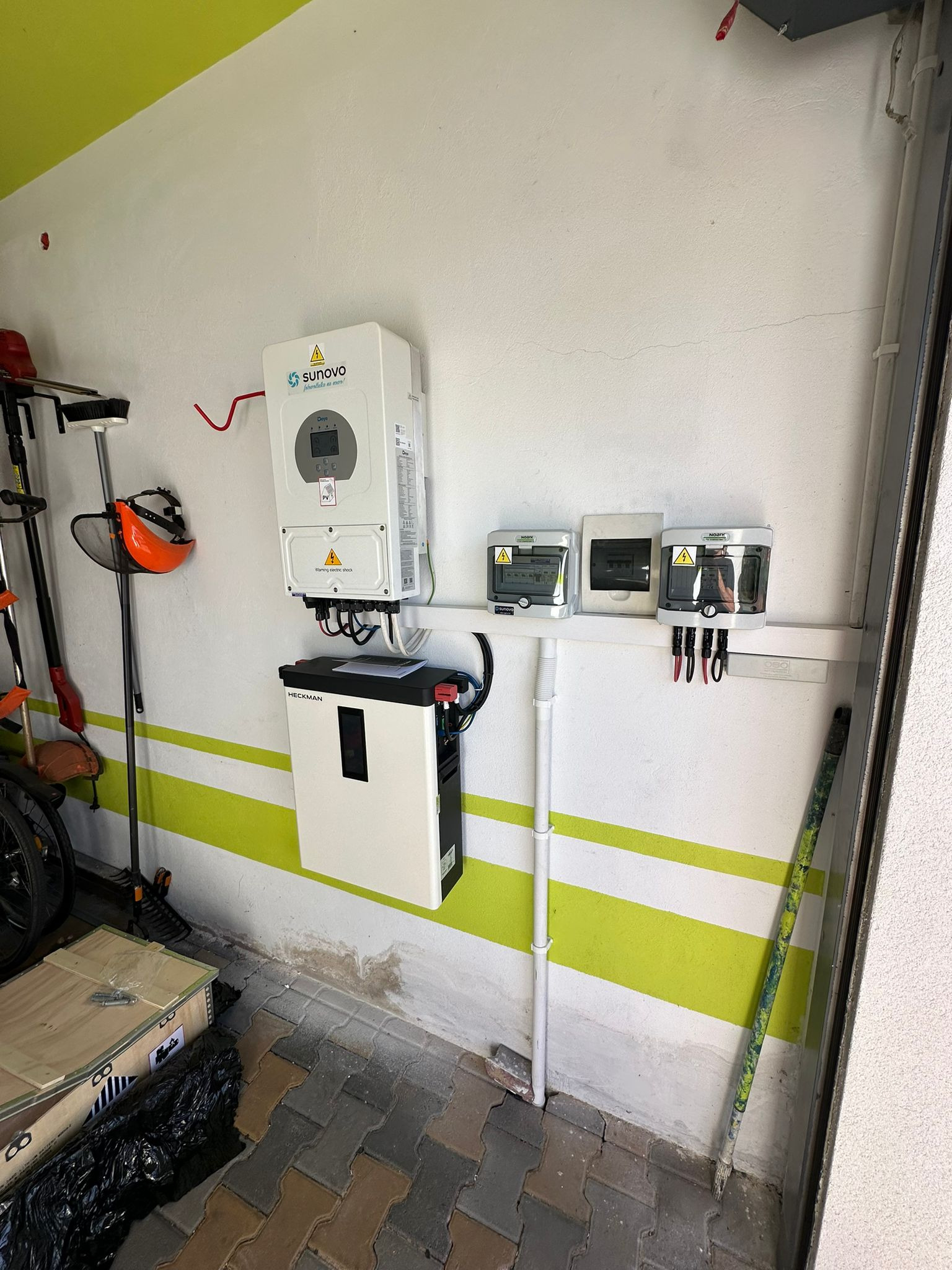 Wnętrze garażu z zamontowanym inwerterem Sunovo, magazynem energii Heckman i zabezpieczeniami, widoczne narzędzia ogrodnicze i skrzynia, ściana w biało-zielone pasy.